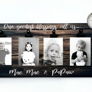 May include: A rustic wooden photo frame with four clips for holding pictures. The frame has the text "Our greatest blessings call us..." and "Mac Mac & Papaw" written on it. The frame is decorated with a woodgrain pattern and has a black border.