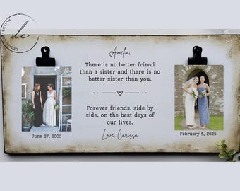 Op de afbeelding: Een rustiek houten bord met twee fotoclips, die elk een foto van twee vrouwen vasthouden. Het bord heeft een handgeschreven boodschap die luidt: "Amelia, er is geen betere vriend dan een zus en er is geen betere zus dan jij. Voor altijd vriendinnen, zij aan zij, op de beste dagen van ons leven. Liefs, Carissa". De data "27 juni 2000" en "5 februari 2025" zijn onder de foto's gedrukt.