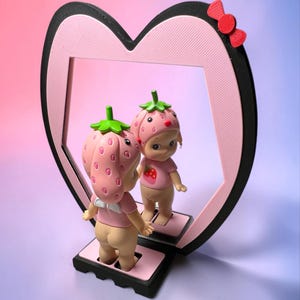 Puede incluir: Un espejo en forma de corazón con marco negro y borde interior rosa, con un pequeño lazo rojo en la parte superior derecha. Una pequeña figura con gorro y camiseta de fresa rosa se encuentra sobre la base, frente a su propio reflejo.
