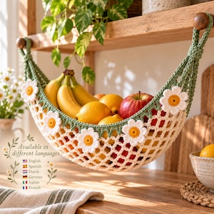 Puede incluir: Una hamaca para frutas tejida a ganchillo en color verde salvia con aplicaciones de margaritas blancas y centros amarillos. La hamaca está suspendida por dos pomos de madera y contiene plátanos amarillos, limones y una manzana roja. El texto dice: Available in different languages.