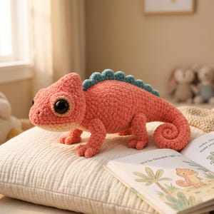 May include: A crocheted chameleon plush toy featuring a textured coral-pink body, a light beige underbelly, and a teal-colored serrated crest along its back. The creature has large, glossy black eyes and a curled tail, crafted from soft yarn.