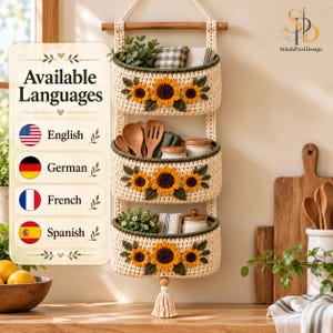 Op de afbeelding: Een hangende gehaakte opbergmand met drie niveaus in crème met donkergroene rand, voorzien van zonnebloemapplicaties op elke mand. De unit hangt aan een houten stok en heeft een kwastje. De tekst luidt Available Languages.