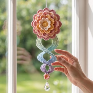 May include: A hanging crochet decoration featuring a multi-layered flower in shades of cream, yellow, orange, and pink. Below the flower is a spiralling, gradient-coloured crochet tail transitioning from green to blue to purple, ending in a crystal pendant.