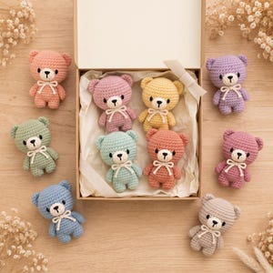 Op de afbeelding: Een verzameling kleine, gehaakte teddyberen in diverse pastelkleuren, waaronder roze, geel, blauw, groen en paars. Elke beer heeft een wit geborduurde snuit, zwarte ogen en een klein wit lintje dat om de nek is gestrikt.