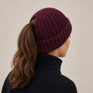 May include: A deep burgundy, ribbed knit beanie featuring a circular opening at the crown designed to accommodate a ponytail. The hat has a thick, folded brim and a textured, vertical stitch pattern that covers the entire surface of the cap.