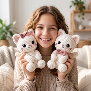 Op de afbeelding: Twee gehaakte kattenknuffels in wit met een zachte garentextuur. Eén kat heeft een klein roze strikje op het oor, terwijl de andere een roze strikje bij de nek heeft. Beide katten hebben grote zwarte ogen, geborduurde roze neuzen en snorharen.