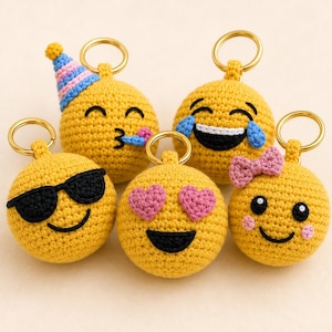 May include: Five round, yellow, crocheted emoji keychains with gold-tone metal rings. One wears a blue and pink party hat, one has black sunglasses, one has pink heart eyes, one has blue tears of joy, and one has a pink bow and rosy cheeks.