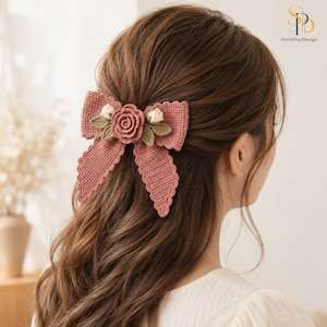 May include: A dusty rose crochet hair bow featuring a central rose motif, two small cream-colored rosebuds, and green crocheted leaves. The bow has a scalloped edge design and is attached to the back of brown hair, showcasing intricate yarn texture.