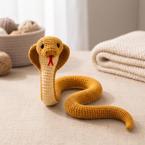 May include: A mustard yellow and cream-coloured crochet cobra toy with a flared hood, black plastic eyes, and a small red yarn tongue. The snake is crafted with a textured stitch pattern and is positioned in an upright, coiled posture.