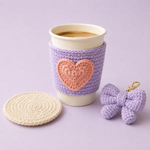 May include: A white paper coffee cup featuring a lavender crochet sleeve with a peach-colored heart appliqué, accompanied by a round cream-colored crochet coaster and a lavender crochet bow keychain with a gold-toned metal clasp.