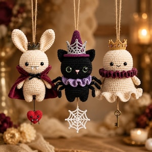 May include: Three hanging crochet ornaments: a cream-colored rabbit with a dark red cape and a small heart, a black spider with a purple witch hat and a white web, and a cream-colored ghost with a purple ruffled collar and a gold key.