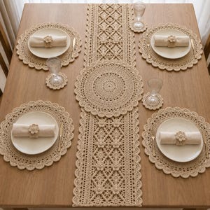 Puede incluir: Un camino de mesa de ganchillo beige con un patrón de diamantes y flores, acompañado de cuatro manteles individuales redondos a juego, cuatro posavasos redondos y cuatro servilletas de tela beige sujetas con servilleteros de flores.