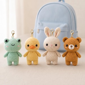 May include: Four crocheted animal keychains featuring a light green frog, a yellow duck, a beige rabbit, and a brown bear. Each figure has black bead eyes, pink stitched cheeks, and a gold-toned metal lobster clasp attached to the top of the head.