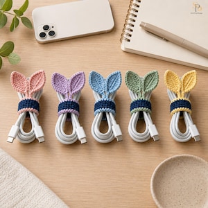 May include: Five crochet cable organizers in pink, purple, blue, green, and yellow. Each organizer features two leaf-shaped accents and a dark blue band that secures a white charging cable. The items are arranged in a row on a wooden surface.