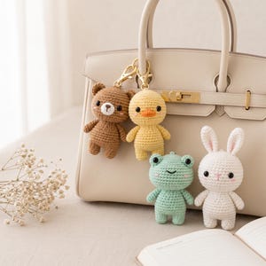 May include: A collection of four small crochet animal keychains including a brown bear, a yellow duck, a light green frog, and a white rabbit. The bear and duck feature gold-toned metal lobster clasps attached to the handle of a beige handbag.