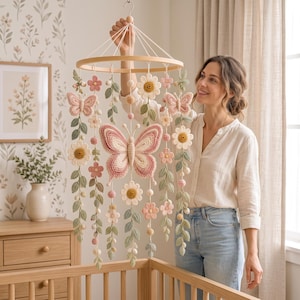 May include: A hanging mobile featuring a circular wooden frame with crocheted elements including a large central butterfly, smaller butterflies, various flowers, and green leaf vines in shades of pink, cream, and sage green, suspended by white strings.