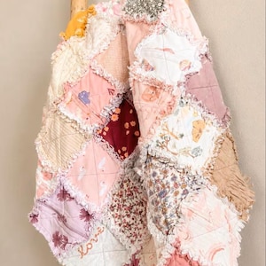 May include: A patchwork rag quilt featuring a variety of fabric squares in shades of pink, white, tan, and deep red. The quilt displays diverse patterns including floral prints, solid colors, and small illustrations. The word love is stitched in cursive.