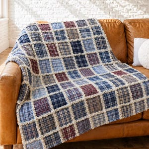 May include: A rag quilt featuring a grid of square fabric patches in various shades of blue, white, and maroon plaid patterns. The squares are joined with exposed, frayed cream-coloured seams, creating a textured, dimensional grid across the surface.