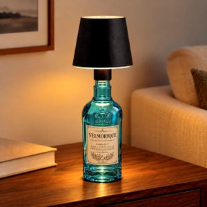 May include: A table lamp constructed from a teal glass bottle base with a black tapered lampshade. The bottle features a cream-colored label with the text Maison Velmorique Collection Botanique Edition No. 07, Aurel, Vesna, Olive, Ornements & Tradition.