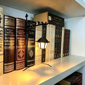May include: A black metal miniature street lamp bookend with a glowing light, positioned between a row of hardcover books on a shelf. The lamp features a classic lantern design with a decorative bracket and emits a warm, soft light.