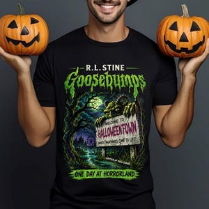 May include: A black t-shirt featuring a graphic print with the text R.L. STINE GOOSEBUMPS, WELCOME TO HALLOWEENTOWN WHERE NIGHTMARES COME TO LIFE, ENTER IF YOU DARE, and ONE DAY AT HORRORLAND. The design includes a green monster, trees, and a sign.