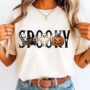 May include: A cream-colored short-sleeve t-shirt featuring the black distressed text SPOOKY. The design includes a black pickup truck carrying orange pumpkins, a white ghost, and an orange jack-o'-lantern with a carved face and black eyes.