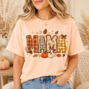 Peut inclure: Un t-shirt à manches courtes couleur pêche arborant le mot MAMA en grandes lettres majuscules à motifs. Chaque lettre présente un design automnal différent, incluant des carreaux et des fleurs, entouré de citrouilles, glands et feuilles.
