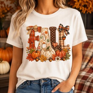 Op de afbeelding: Een wit t-shirt met korte mouwen en een afbeelding van het woord FALL in grote, textuurrijke letters. De letters zijn versierd met ruitjes, stippen en bladpatronen, geaccentueerd door strikken, pompoenen, eikels en kleurrijke herfstbladeren.
