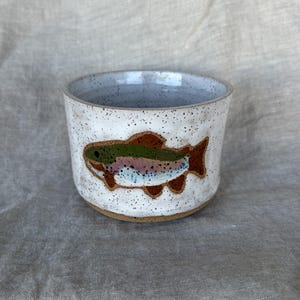 May include: A ceramic cup with a speckled white glaze and a textured surface. The exterior features a hand-painted illustration of a trout with green, pink, and brown markings. The interior of the cup is a smooth, light grey speckled finish.