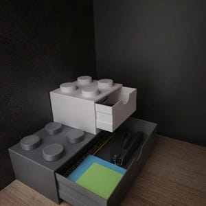 Può includere: Un'unità di archiviazione modulare bianca e grigia a forma di mattoncini da costruzione. L'unità bianca presenta un cassetto estraibile, mentre la base grigia più grande contiene un cassetto largo con una cucitrice nera, matite e foglietti adesivi.