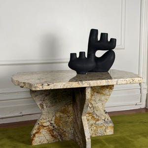 May include: A rectangular granite table with a polished top and a cross-shaped base featuring mottled beige, brown, and black stone patterns. Two matte black ceramic sculptures with multiple rounded, finger-like protrusions sit on the table.