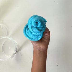 May include: A bright cyan, pliable, and glossy substance with a swirled, textured surface. The material is shaped into a rounded, coiled mound. Beside the substance sits a clear, empty, circular plastic container with a matching lid.