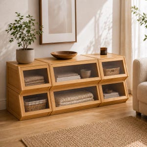 May include: A modular wooden storage unit consisting of six stacked compartments with angled front frames and clear glass panels. The light-toned wood structure features open shelving for organizing folded textiles, woven baskets, and books.