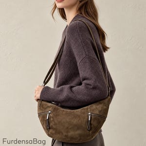 May include: A dark olive green suede hobo bag featuring a curved silhouette, an adjustable shoulder strap, and two front zippered pockets with silver-toned hardware. The text "FurdensaBag" is printed in the bottom left corner of the image.