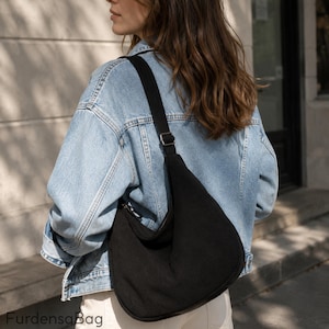 Puede incluir: Un bolso de hombro negro estilo hobo con una superficie de tela texturizada y una correa negra ancha y ajustable con una hebilla de metal plateado. El bolso se lleva al hombro y el texto FurdensaBag es visible en la esquina inferior izquierda.
