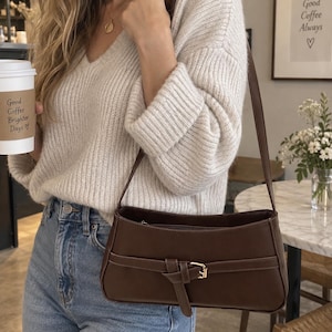 May include: A dark brown shoulder bag with a rectangular silhouette, featuring a decorative horizontal strap across the front secured by a gold-toned metal buckle. The bag has a single shoulder strap and a top zipper closure.