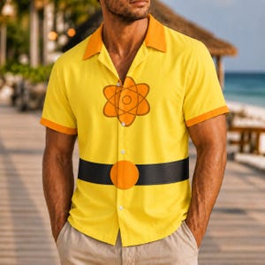 Op de afbeelding: Een felgeel overhemd met korte mouwen en knoopsluiting, voorzien van een oranje atoom-symbool op de borst en een zwarte riem-print rond de taille met een grote ronde oranje gesp. Het overhemd heeft een oranje kraag en mouwboorden.
