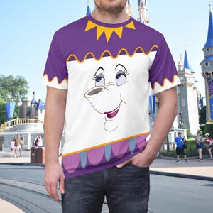 May include: A short-sleeved t-shirt featuring a graphic print of the character Mrs. Potts from Beauty and the Beast. The shirt has a purple top section with a yellow sun-like collar, a white center, and a purple and blue scalloped hem design.