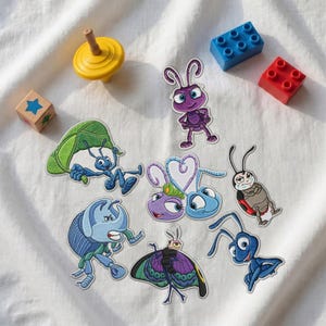 Può includere: Una collezione di toppe ricamate raffiguranti vari personaggi insetti del film A Bug's Life, tra cui figure blu, viola e rosse, insieme a una trottola gialla, un blocco di legno con una stella blu e due mattoncini di plastica.