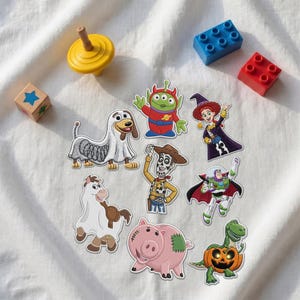 May include: A collection of embroidered patches featuring characters from Toy Story in Halloween costumes, including a slinky dog, a green alien, a witch, a skeleton cowboy, a space ranger with a cape, a horse, a pig, and a dinosaur in a pumpkin.