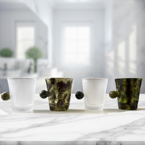 May include: Four small glass cups arranged in a row. Two cups are made of frosted white glass, and two cups feature a mottled, dark green and black textured pattern. Each cup includes a single, small, round ball-shaped handle on the side.