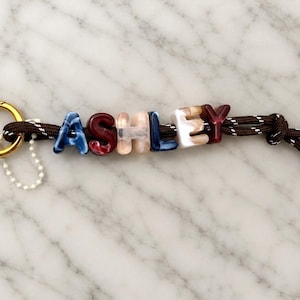 May include: A keychain featuring a gold-toned metal ring attached to a brown cord with white flecks. The cord is threaded through acrylic letter beads spelling ASHLEY in blue, red, peach, and white marbled colors, with a small white bead.