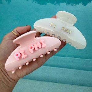 May include: Two hair claw clips, one in light pink with raised pink lettering reading PI PHI and one in translucent white with gold lettering reading PI PHI. Both clips feature a curved design with multiple teeth for securing hair in place.