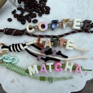 May include: Three wristlet keychains made of nylon cord with gold-tone metal rings. The top keychain features brown cord and beads spelling COFFEE. The middle keychain has black and white cord and beads spelling LATTE. The bottom keychain has light green cord and beads spelling MATCHA.