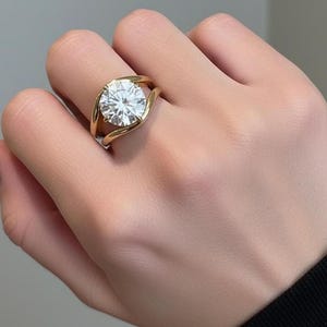 May include: A gold ring featuring a large, round, brilliant-cut clear gemstone set in a bypass-style band. The band curves around the central stone, creating a smooth, continuous gold frame that highlights the faceted clarity of the centre gem.