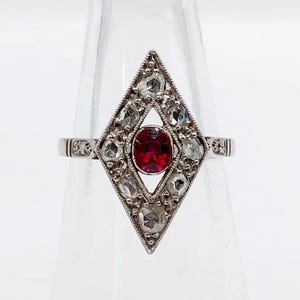 May include: A silver-toned ring featuring a central oval-shaped red gemstone set within a diamond-shaped frame. The frame is decorated with multiple small, clear, faceted stones arranged in a geometric pattern around the center stone.