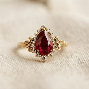 Peut inclure: Une bague en or avec une pierre précieuse rouge foncé en forme de poire au centre, entourée d'un halo de petites pierres blanches. L'anneau présente des détails de volutes complexes des deux côtés, partant du sertissage central.