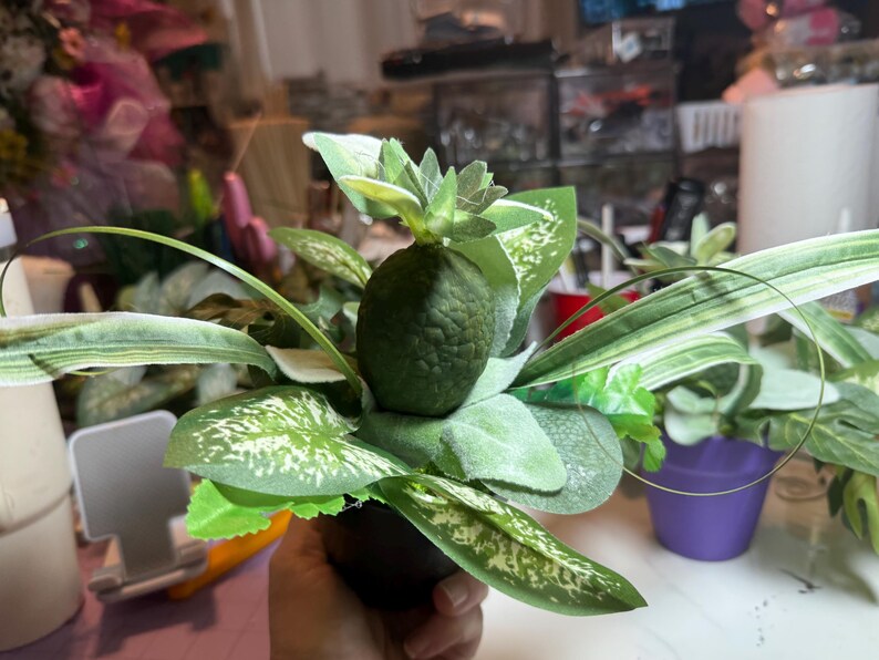 Op de afbeelding: Een decoratief kunstplantarrangement met een centraal donkergroen, getextureerd element in de vorm van een ananas, omringd door diverse groene bladeren, waaronder enkele met witte spikkels en lange, dunne, puntige bladeren in een donkere pot.