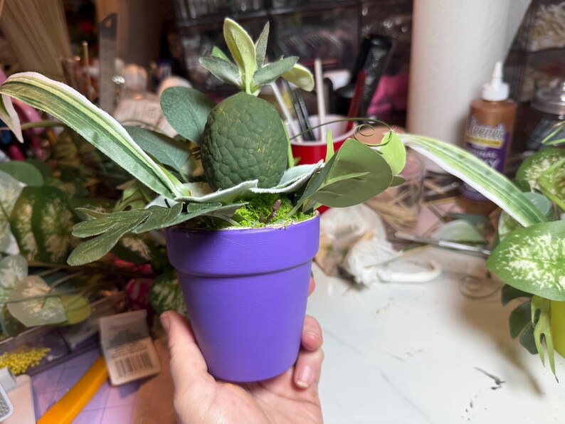 Op de afbeelding: Een kleine paarse bloempot met een getextureerde, donkergroene, ovaalvormige vrucht of decoratief element, omringd door diverse groene kunstbladeren en gebladerte. Het plantarrangement staat in de effen paarse pot.