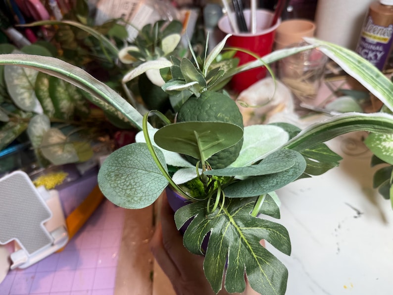 Op de afbeelding: Een decoratief arrangement van kunstgroen met een centrale donkergroene, getextureerde vrucht, omringd door diverse bladvormen, waaronder lange, bonte sprietbladeren, afgerond eucalyptusachtig blad en een donkergroen monstera-blad.